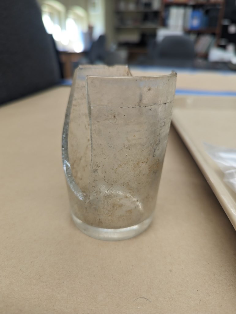 a picture of a broken glass jar on a laboratory table with a sorting tray beside it. The jar has five rows of small vertical lines which start about 1.5 cm below the rim.